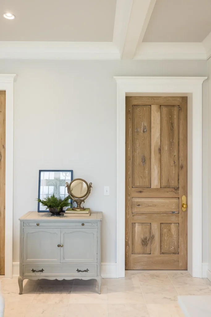 A light wood door with a gold handle is set in a white-trimmed wall beside a vintage light gray cabinet topped with a round mirror, a framed photo, and a small green plant on a marble tile floor.