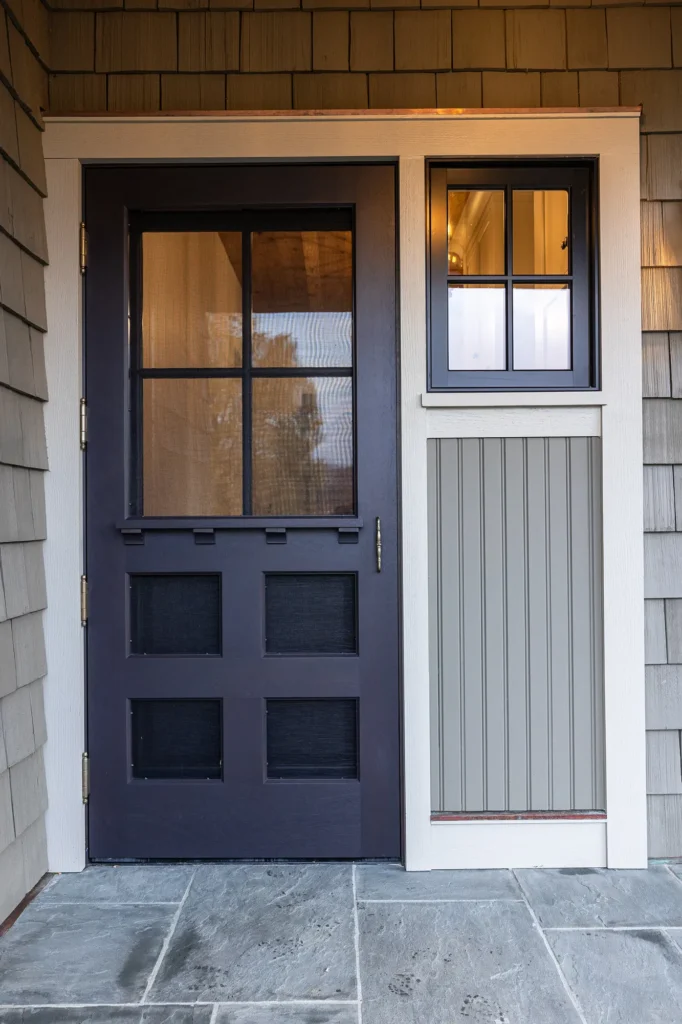 A front door with four glass panels and a black frame next to a small window with matching trim, set in a house with gray siding and stone tile flooring.