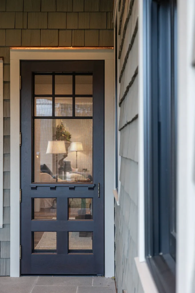 A dark blue screen door with a grid window design opens to a cozy interior with a lit table lamp and a wreath visible through the mesh. The house exterior has gray shingles and white trim.