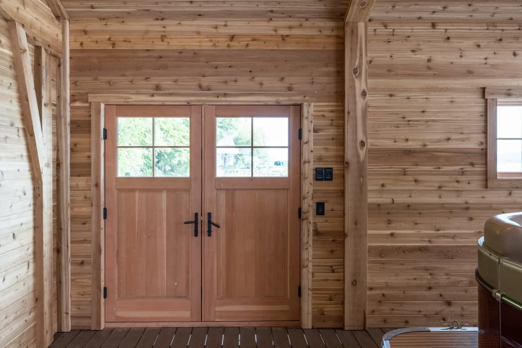 Double wooden doors with glass panes and black handles are set in a wall of light-colored wooden planks. Sunlight and greenery are visible through the door’s upper windows. A small window and light switches are on the right.