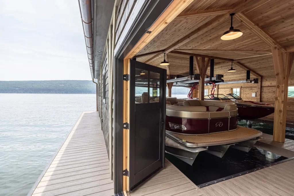 A modern boathouse with wooden walls and ceiling, open sliding door, and two docked pontoon boats inside, sits on a calm lake next to a long, narrow boardwalk. Forested hills are visible in the distance.