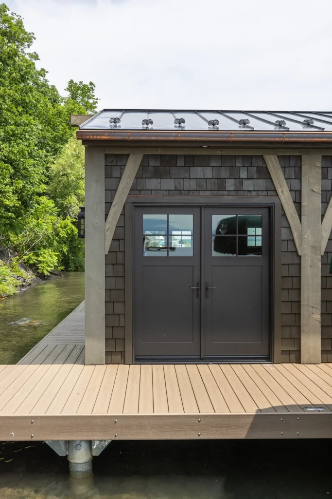 A dark-shingled boathouse with double glass doors sits on a wooden dock over clear water, surrounded by lush green trees under a partly cloudy sky.