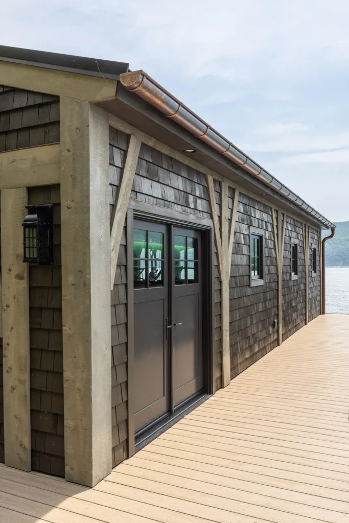 A rustic lakeside building with gray shingles and large wooden beams sits beside a wide, light-colored wooden deck overlooking calm water under a cloudy sky.