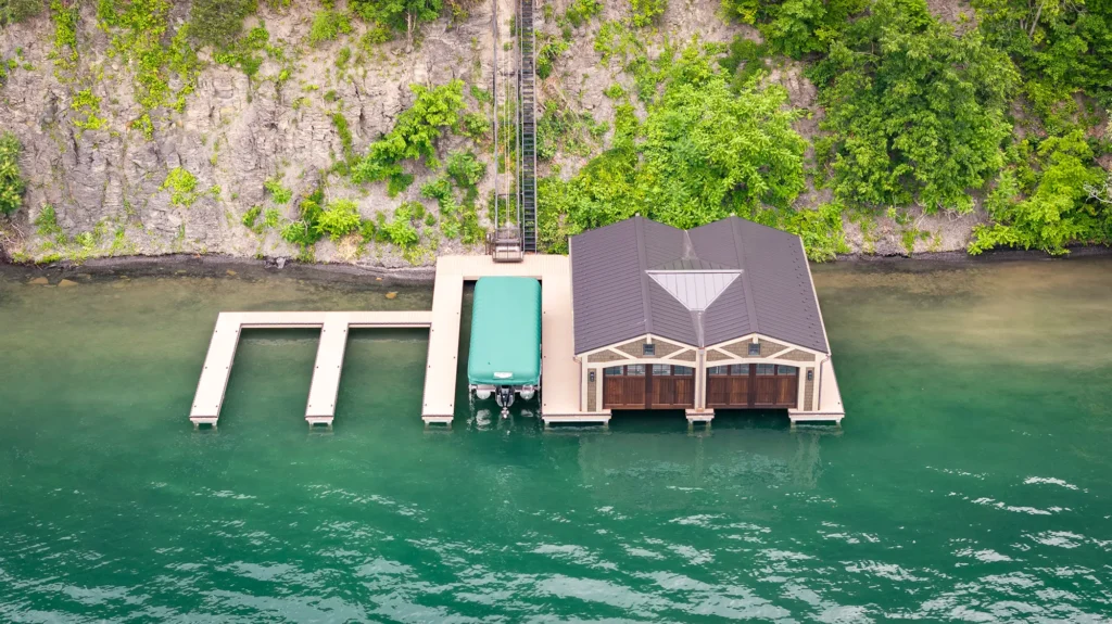 A boathouse with a dock and covered boat slip sits on clear turquoise water at the base of a rocky, tree-covered hillside, with a rail track running up the hill.