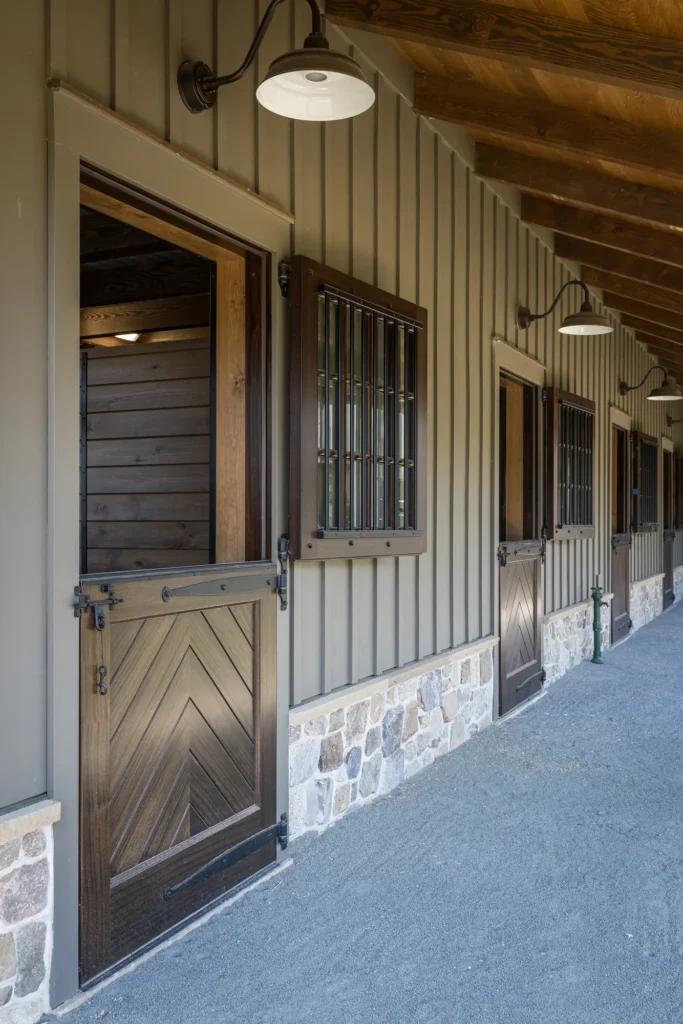 A row of wooden stable doors with metal bars and stone accents, under a covered walkway with hanging lights. The area appears clean and well-maintained.