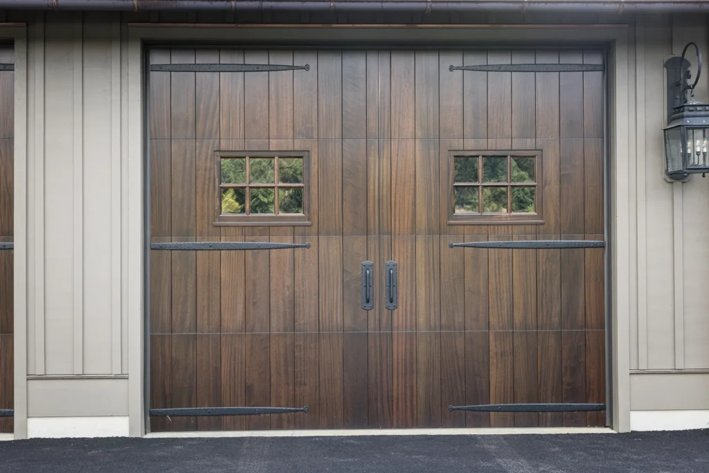 A wooden double garage door with two small square windows, black iron hinges, and handles. The door is set in a beige exterior wall with a black lantern mounted on the right side.