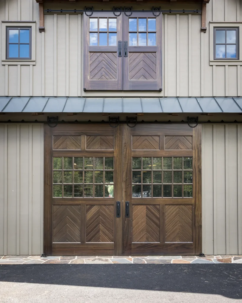 Two large wooden garage doors with chevron patterns and glass panes stand below matching double windows on a beige vertical-paneled building, featuring black metal handles and rustic trim.