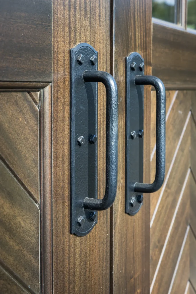 Close-up of two black metal handles on a pair of wooden doors with a chevron pattern. The wood is brown with visible grain, and the handles are attached with black bolts.