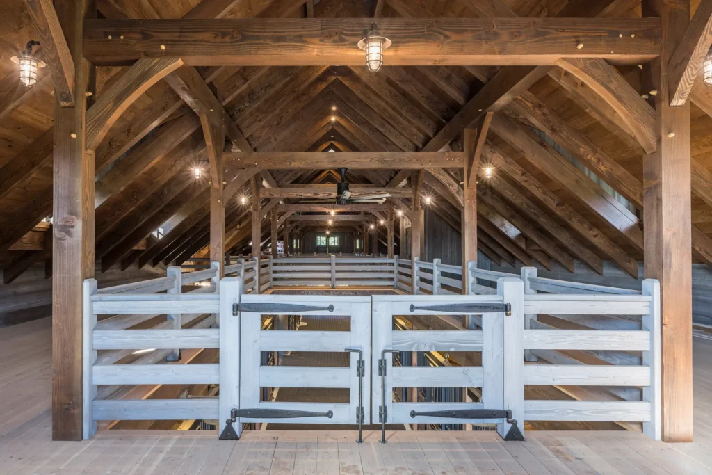 View of a rustic barn interior with exposed wooden beams, warm lighting, and a white wooden gate in the center. The space has a vaulted ceiling and a symmetrical, open design.