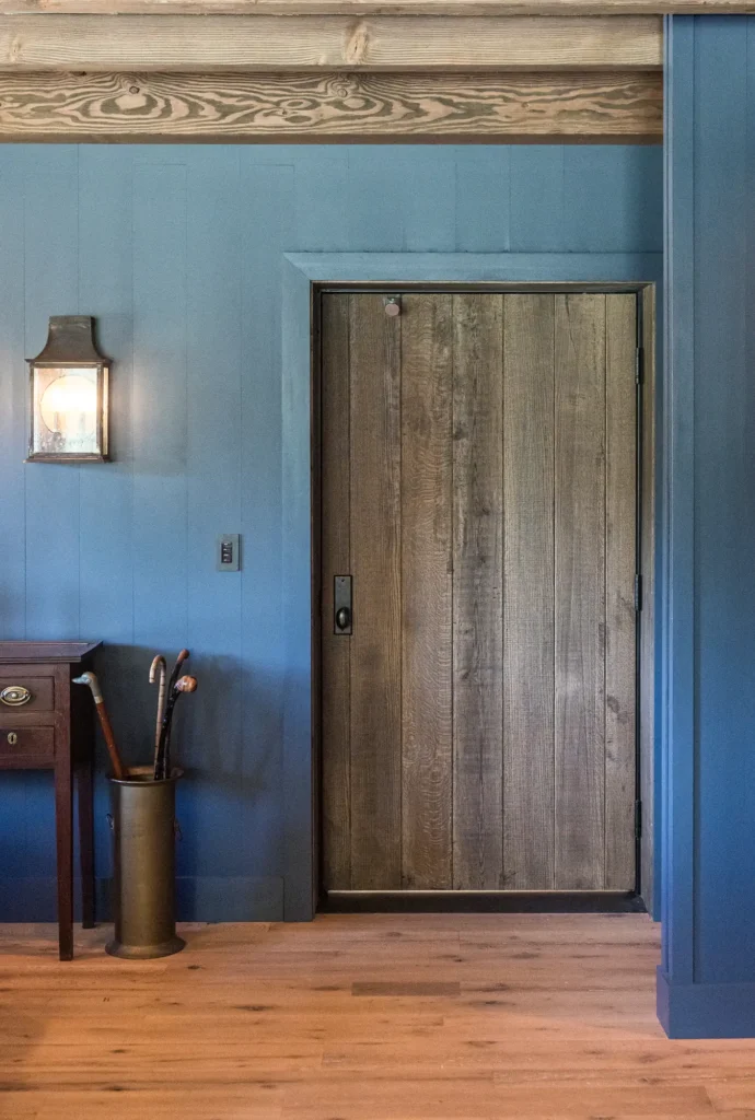 A rustic wooden door set in a blue paneled wall, with a lantern-style light fixture, a cane holder with walking sticks, a small table, and wooden flooring creating a cozy entryway.