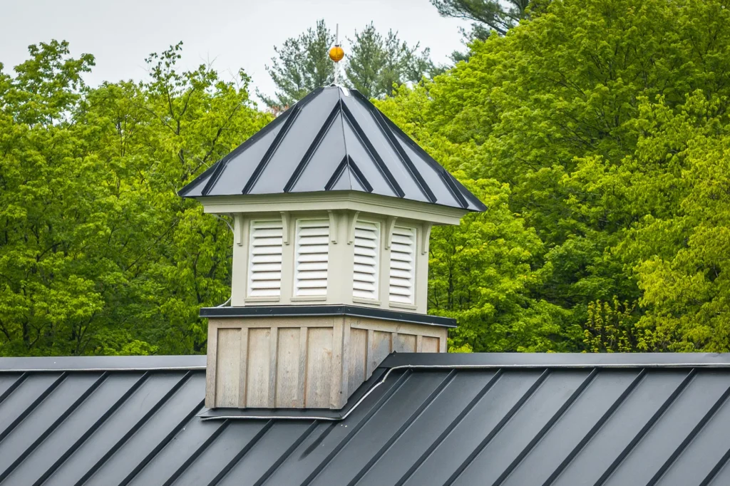 A close-up of a cupola with white louvered vents and a black metal roof, sitting atop a metal-roofed building, surrounded by lush green trees in the background.