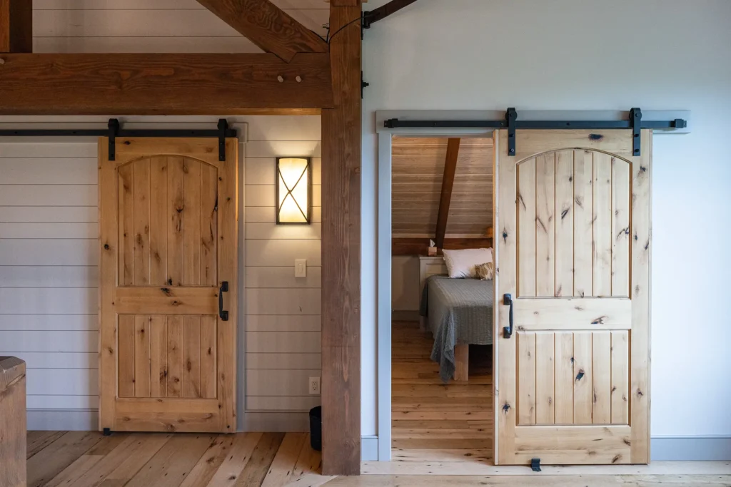 Two wooden sliding barn doors, one closed and one open, reveal a cozy bedroom with a bed inside. The room features wood beams, white shiplap walls, and natural wood floors, creating a rustic, farmhouse atmosphere.