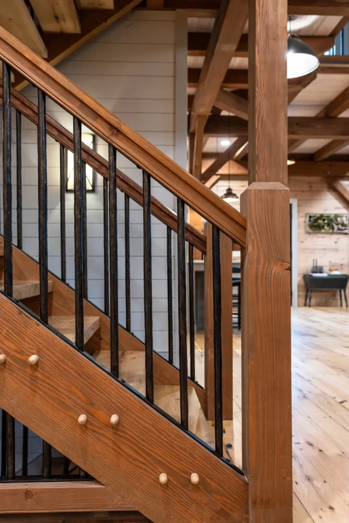 Wooden staircase with black metal balusters in a rustic interior, featuring exposed beams, wood floors, and paneled walls; warm lighting and cozy decor visible in the background.
