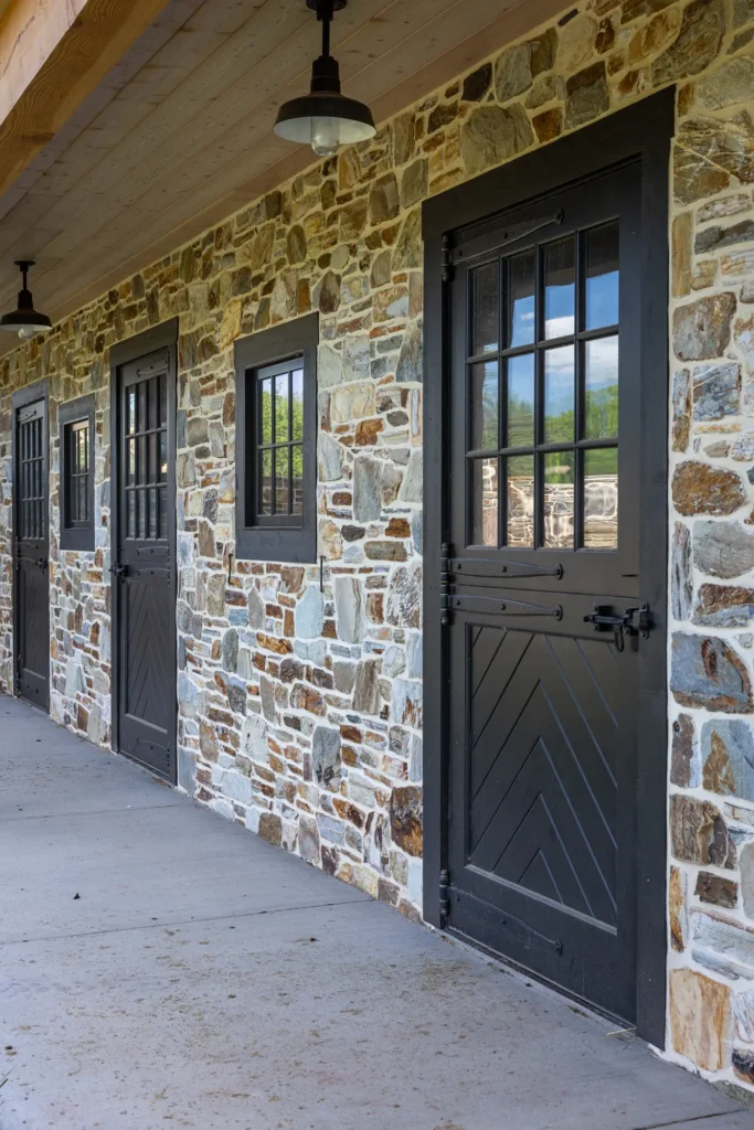 Stone building exterior with three black Dutch doors and matching windows beneath a wooden overhang, illuminated by black industrial-style lights on a concrete walkway.