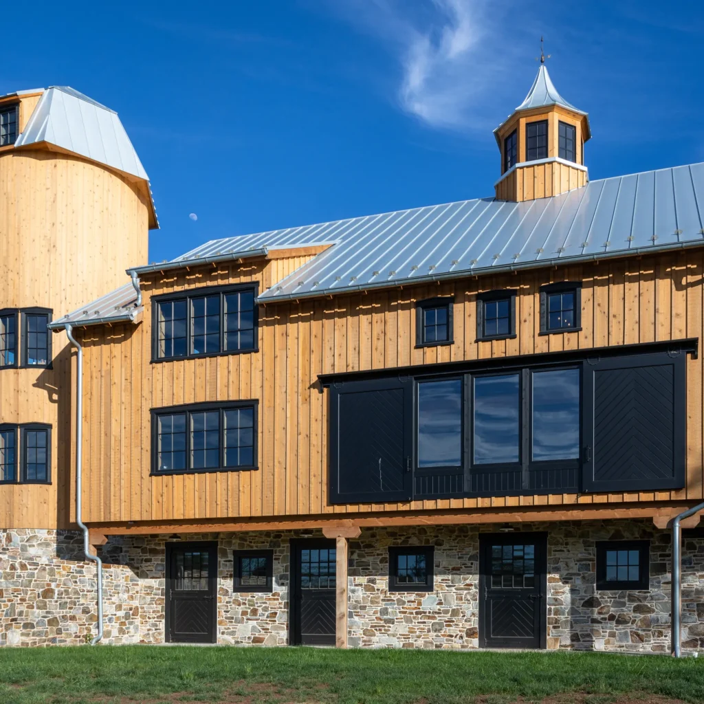 A modern barn with light wood siding, stone foundation, black trim, and a metal roof sits under a clear blue sky with a small moon visible. The barn features large black doors and several windows.