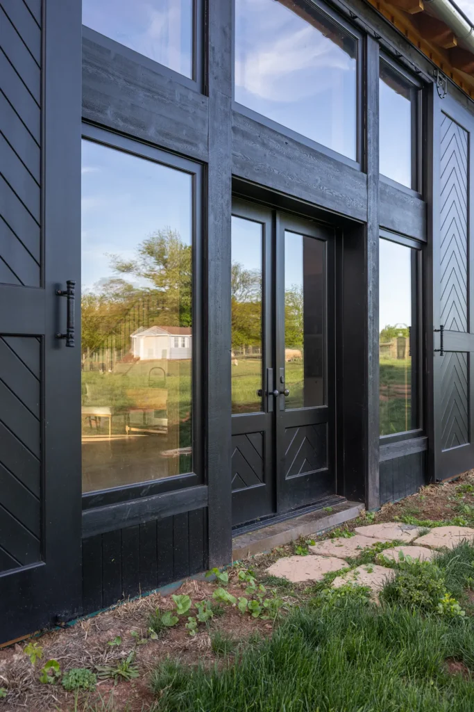 Tall black-framed glass doors and windows on the side of a modern building reflect the outdoor scenery, with grass, plants, and a stone path in the foreground.