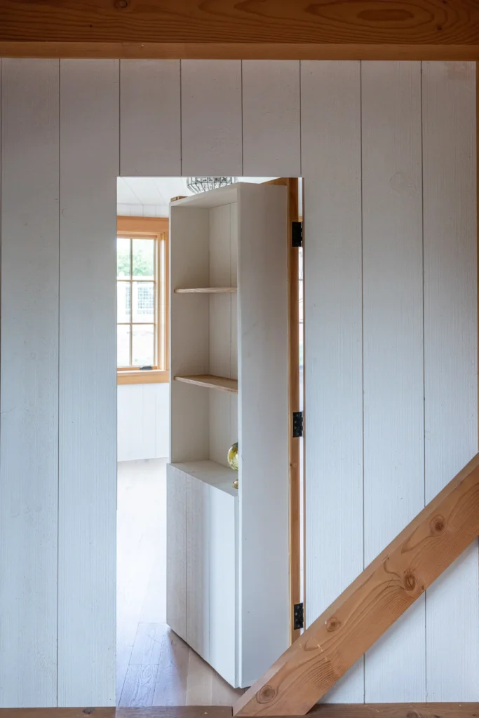 A white built-in shelving unit is cleverly designed as a hidden door, partially open to reveal a bright room with large windows, set in a wall with wooden paneling and a diagonal wood beam.