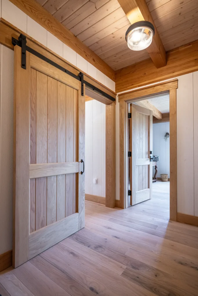 Hallway with light wood floors, wood-paneled ceiling, and wooden sliding barn door on the left; open hinged doors lead to a bright, cozy room. Ceiling light fixture and natural sunlight illuminate the space.
