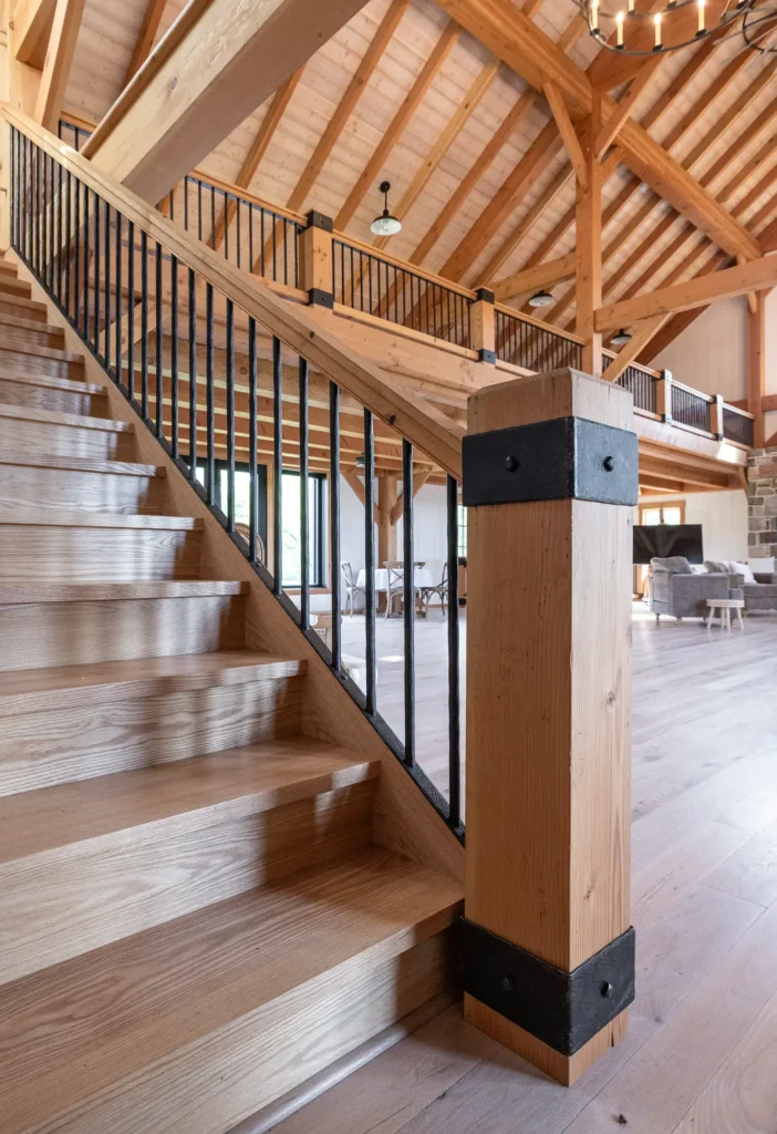 A wooden staircase with black metal railings in a spacious, open-concept room featuring exposed wooden beams, large windows, and modern rustic decor.