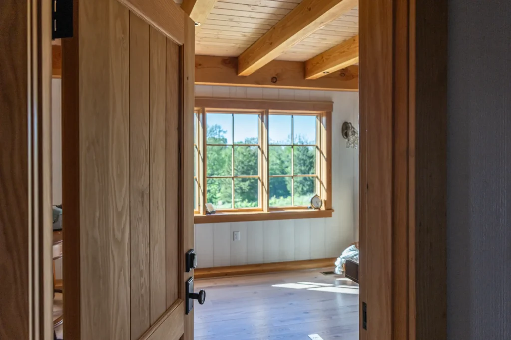 A wooden door opens to a bright room with wooden floors, exposed ceiling beams, and large windows letting in natural light and offering a view of green trees outside.