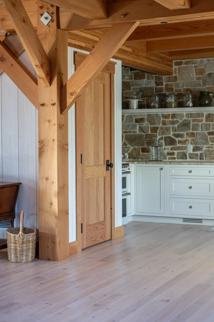 A cozy kitchen with light wood floors, exposed wooden beams, a wooden door, white cabinets, and a stone backsplash. A woven basket sits on the floor near the doorway.