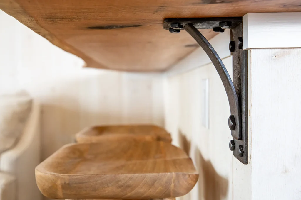 Close-up view of a wooden countertop supported by a black metal bracket, with two wooden bar stools placed underneath against a light-colored wall.