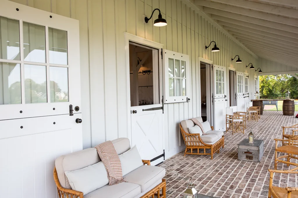 Outdoor patio with wicker chairs and sofas, brick flooring, and white Dutch doors set in a light-colored wooden wall. Black outdoor lights are mounted above each door, creating a cozy, inviting atmosphere.