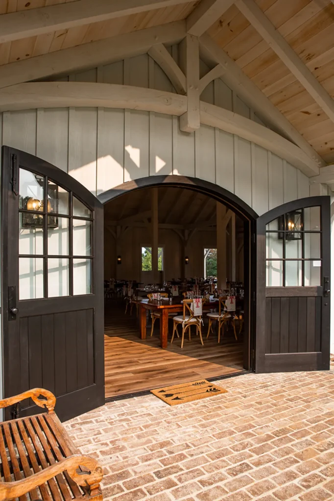 Large black double doors are open, revealing a warmly lit dining area with wooden tables and chairs inside a rustic building with a vaulted wood ceiling and brick floor outside.