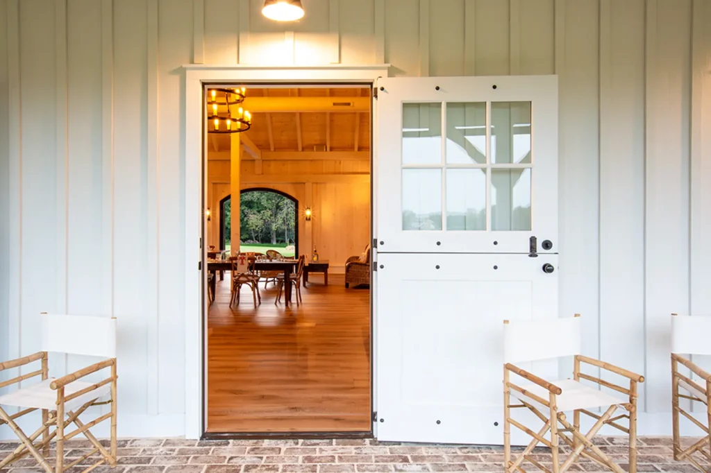 A white Dutch door stands open, revealing a warmly lit interior with wooden floors, tables, and chairs. Two white director’s chairs sit outside on a brick porch against light-colored siding.