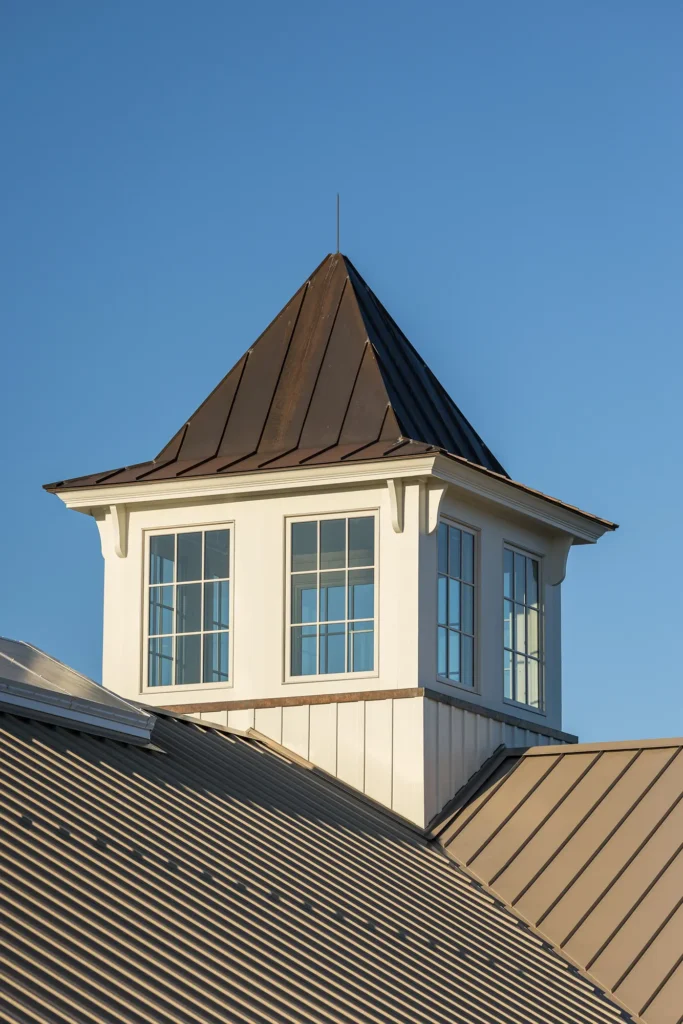 A white cupola with large windows and a brown metal roof sits atop a building with a matching brown metal roof, set against a clear blue sky.