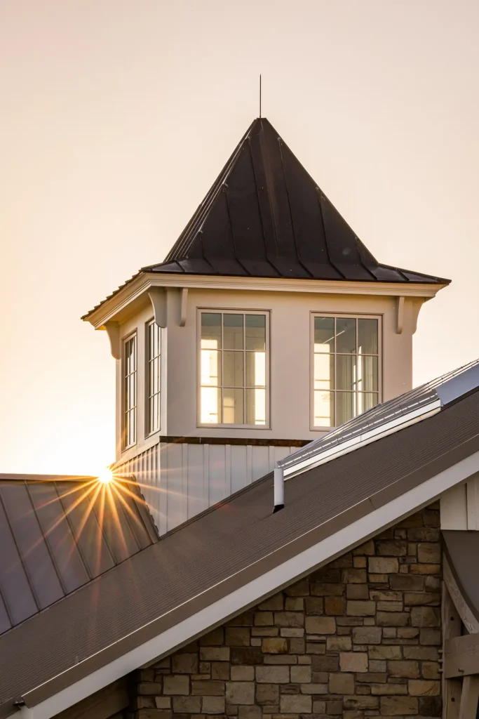 A sunbeam shines through the windows of a white cupola with a dark pointed roof, set atop a building with a stone facade and metal roof at sunset.