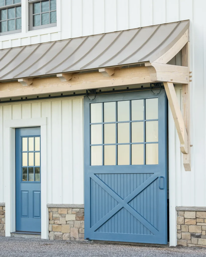 A white building with stone accents features a blue door and a large blue sliding barn door with glass panes and a diagonal cross design. The roof above has a curved metal awning supported by decorative wooden brackets.
