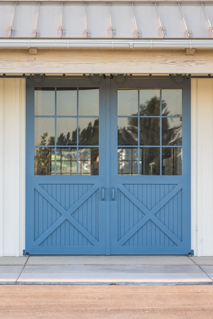 Blue double barn doors with glass window panes and a crisscross wood pattern at the bottom, set in a light-colored building with a metal roof and concrete pathway in front. Trees are reflected in the glass.