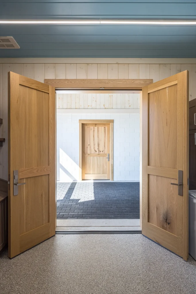 Double wooden doors open to reveal an outdoor area with a black tiled floor, leading to another closed wooden door set in a white wall. The ceiling inside is light blue with a modern light fixture.