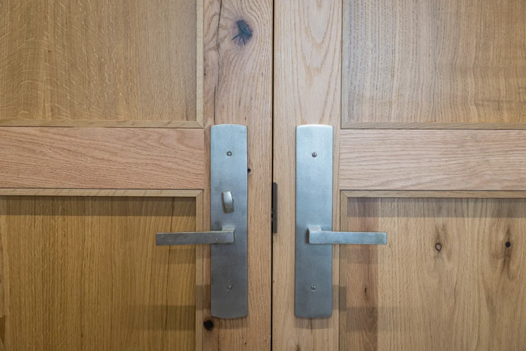 Two wooden doors with metal handles and locks are shown side by side. The wood has a natural finish with visible grain and knots. The handles are rectangular and metallic, positioned horizontally.