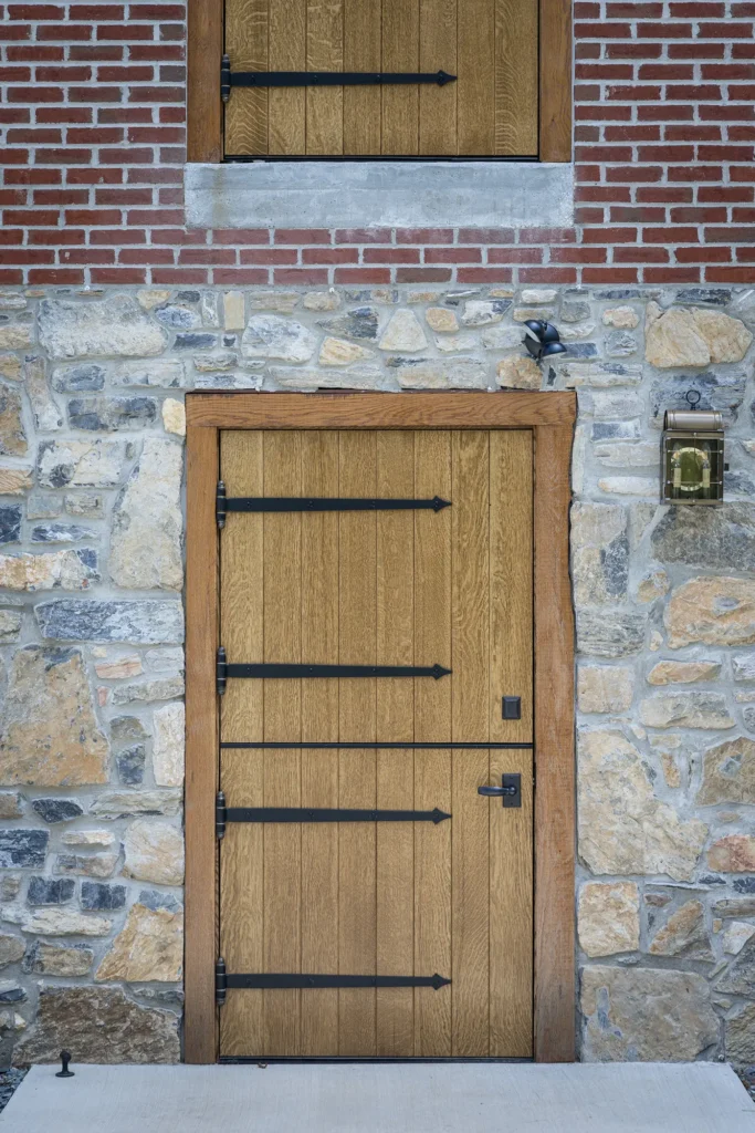 A wooden door with four black metal hinges set in a stone wall, with a brick section above and a lantern mounted on the right side. Another wooden door is visible above the main door.