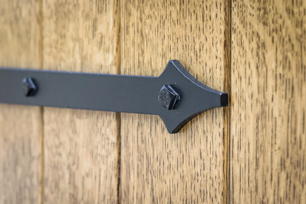 Close-up of a black metal strap hinge with large bolts attached to a wooden door made of vertical planks with a natural wood grain finish.