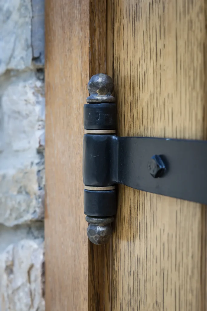 Close-up of a black metal door hinge attached to a wooden door, with a stone wall visible in the background. The wood has a natural grain and the hinge has a slightly worn appearance.