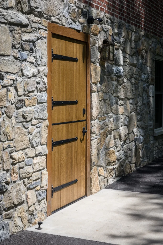 A wooden door with black metal hinges is set in a rustic stone wall. A small outdoor lantern light hangs above the door, and there is a concrete path and some asphalt leading up to the entrance.