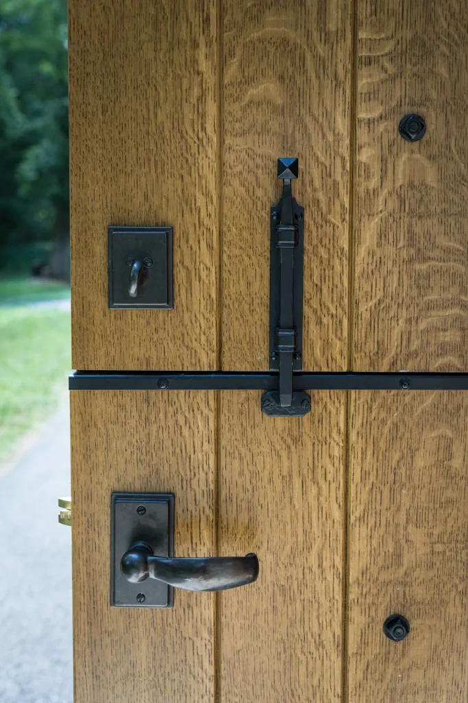 A close-up of a wooden door with vertical panels, featuring a metal handle, a latch lock, and a slide bolt. The door is partially open, revealing greenery and a pathway outside.