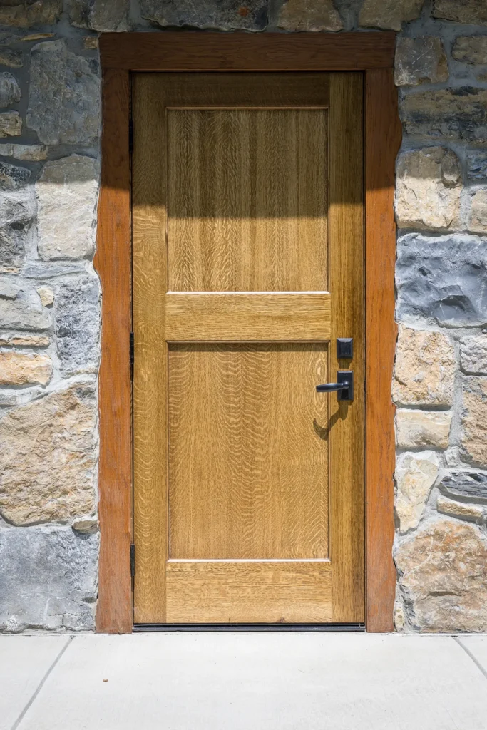 A wooden door with a rectangular design and black handle is set in a stone wall with a concrete sidewalk in front.
