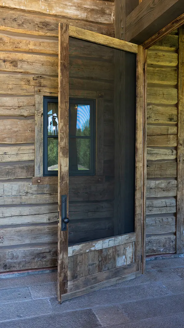A rustic wooden screen door is partially open on a log cabin wall with a small window nearby. The scene has a weathered, natural look and stone flooring.