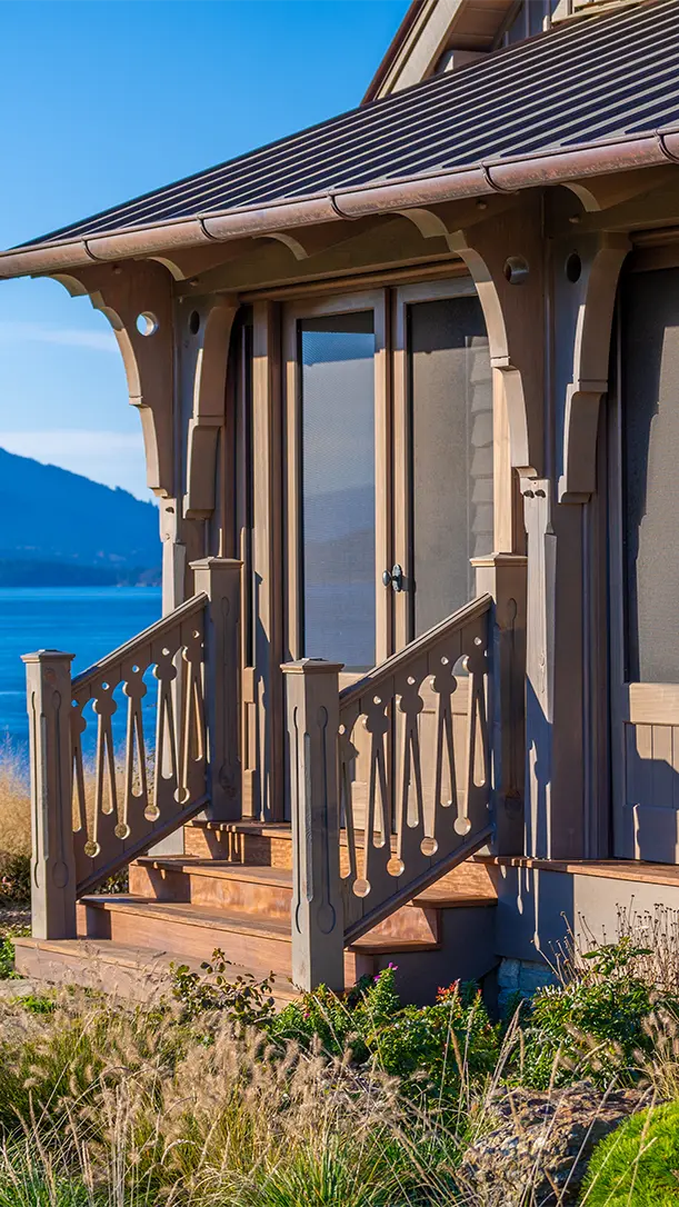 A close-up of a wooden porch with decorative railings and posts, attached to a house overlooking a body of water and distant hills under a clear blue sky.