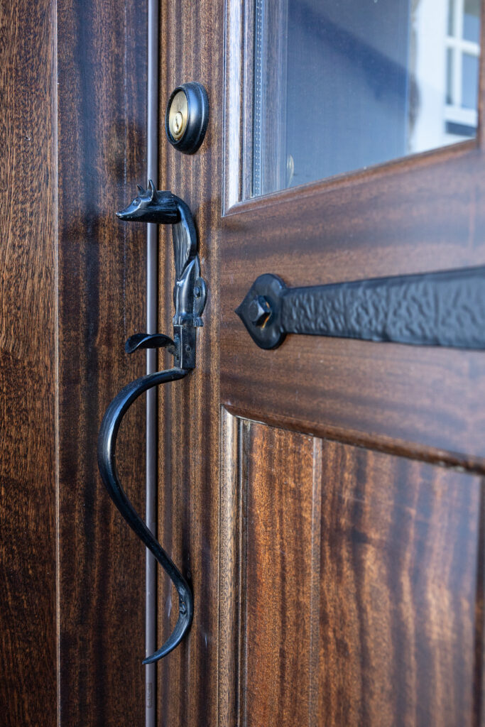 Close-up of a wooden door with a decorative black metal handle and lock. The handle features a curved design, and there is a glass window pane in the upper part of the door.