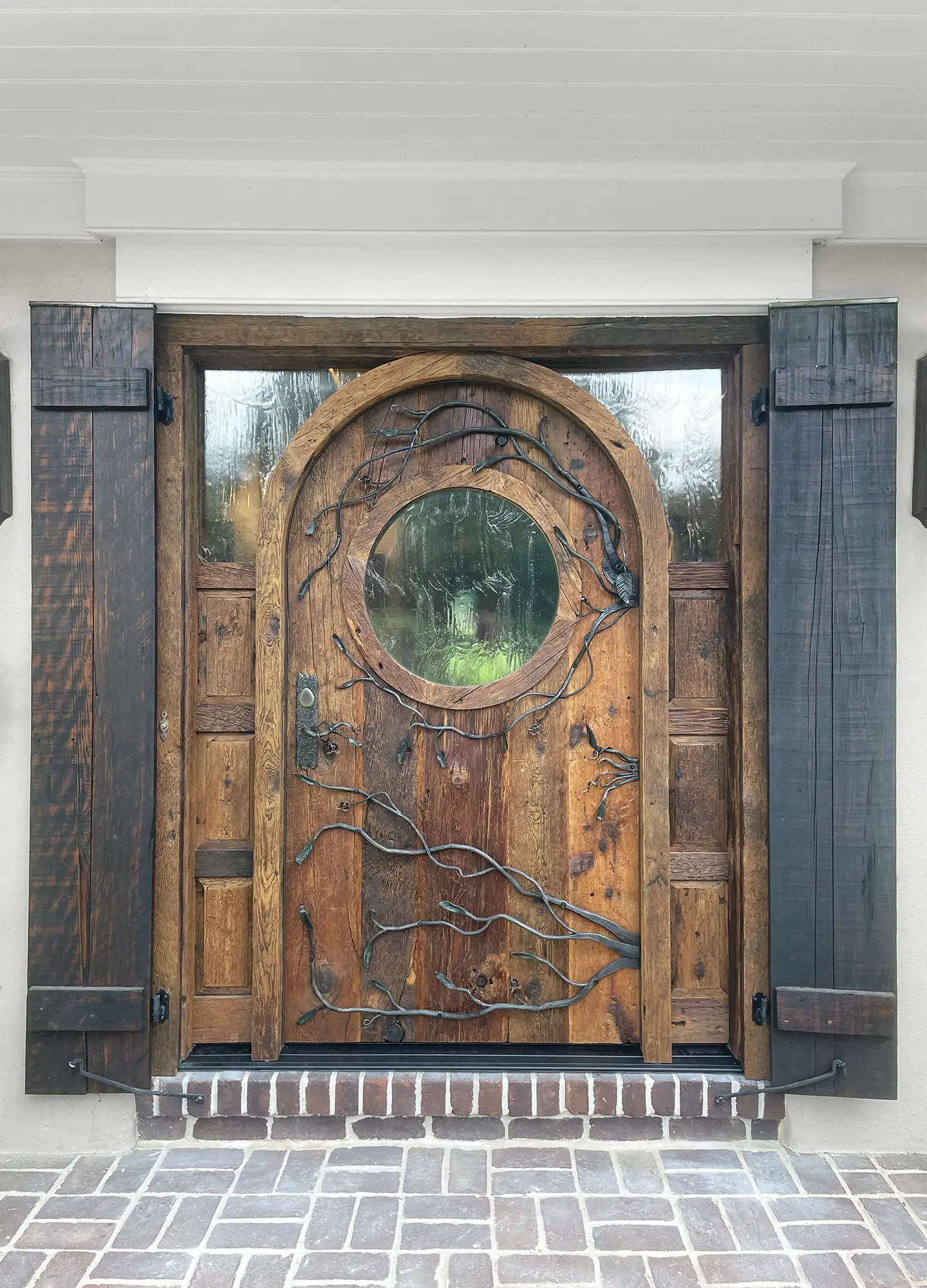 A rustic, arched wooden door with a round window in the center, framed by dark wooden shutters and decorative metal vines, set in a stone and brick entryway.