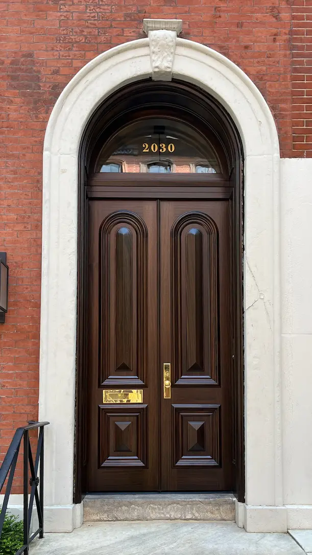 A tall, ornate wooden door with decorative panels and a brass mail slot and handle, set in a white stone arched frame against a red brick wall. The number 2030 is displayed above the door.
