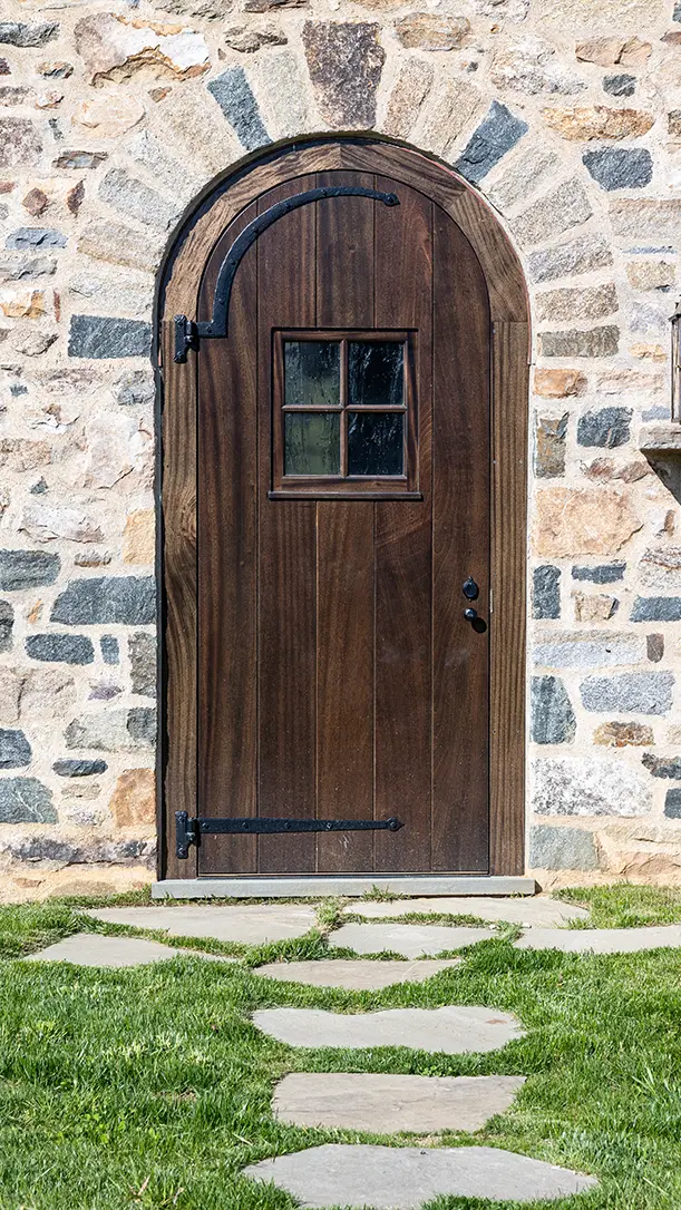 A wooden arched door with a small window and black metal hinges set in a stone wall, with a stone path leading up through the grass.