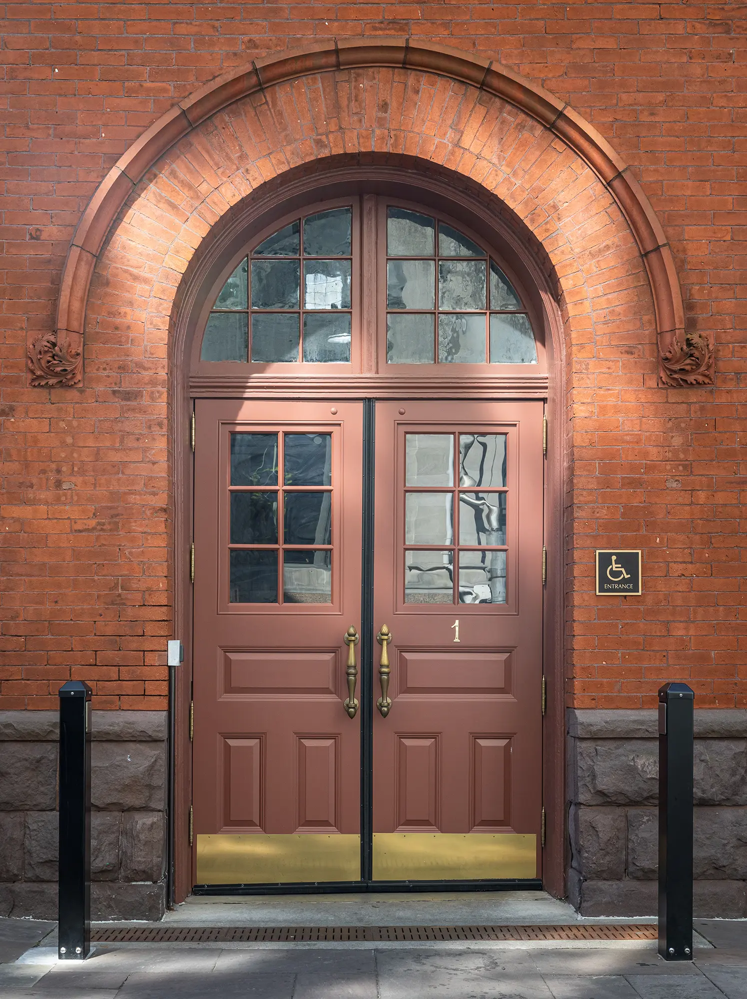 A pair of brown double doors with glass panes set in a red brick building. An arched window is above the doors, and a wheelchair accessibility sign is mounted on the right side of the doorway.
