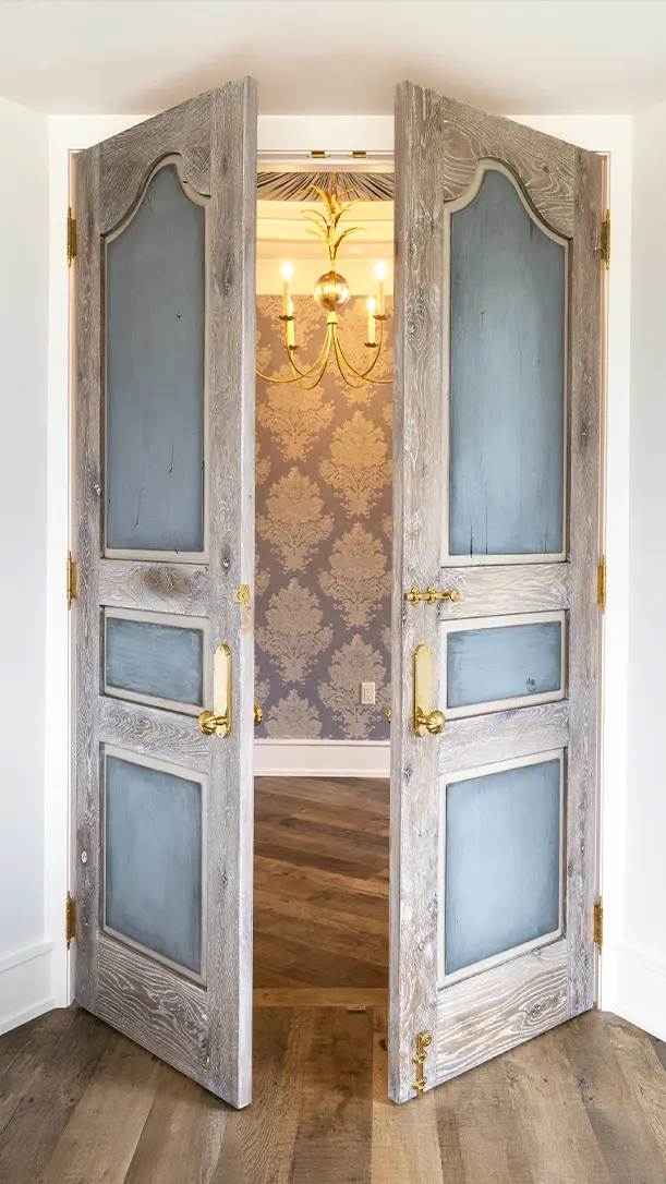 A pair of vintage wooden double doors with blue panels open inward, revealing a small room with damask wallpaper, a wooden floor, and a gold chandelier hanging from the ceiling.