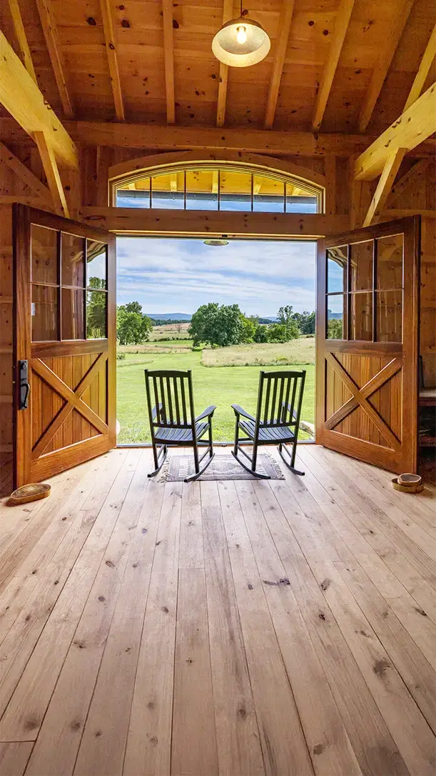 Two wooden rocking chairs face outward through open barn doors, overlooking a green grassy field and distant trees under a partly cloudy sky, inside a rustic wooden barn with exposed beams.